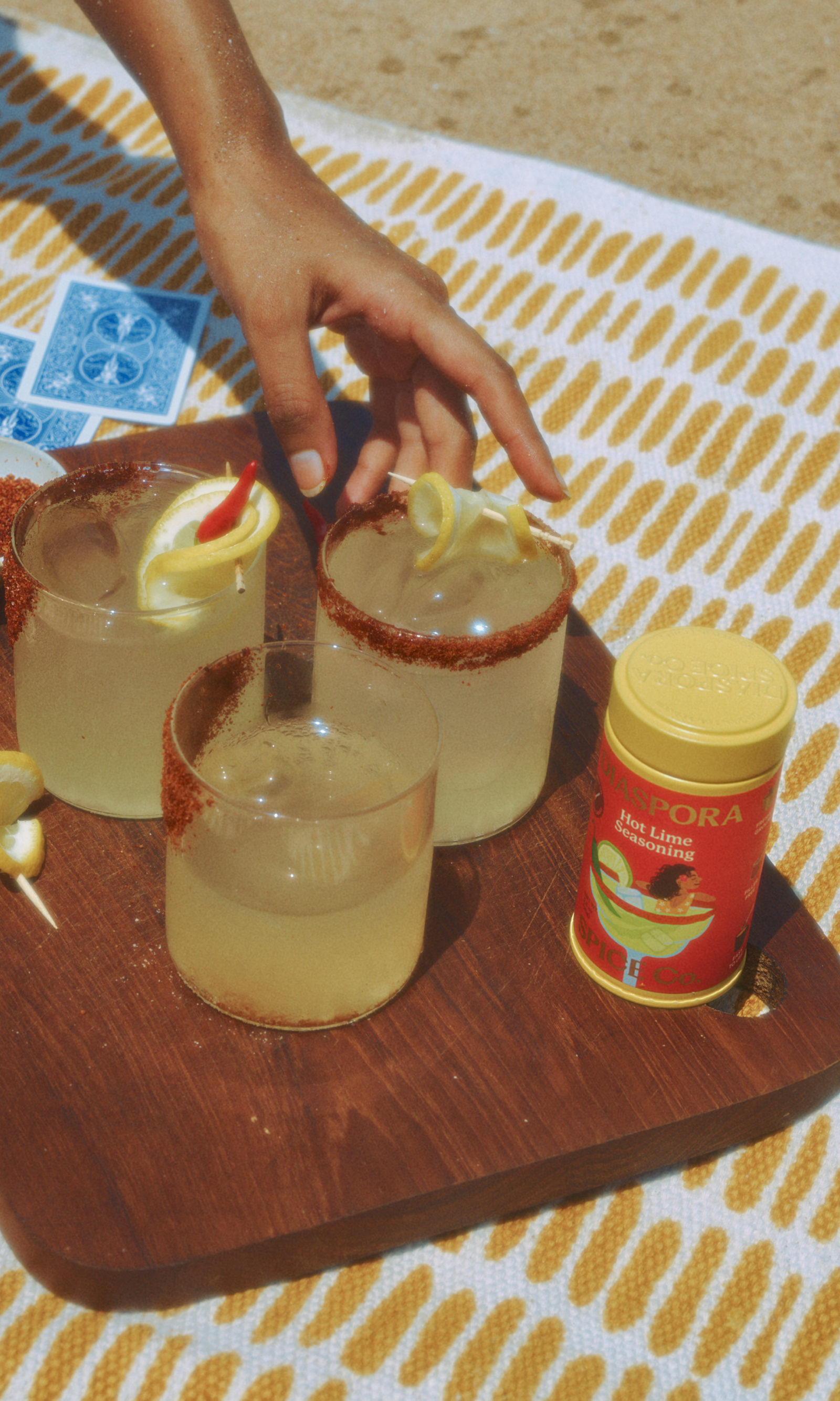 Hot Lim Seasoning tin kept on a picnic blanket along with 2 margarita glasses with a cocktail rim.